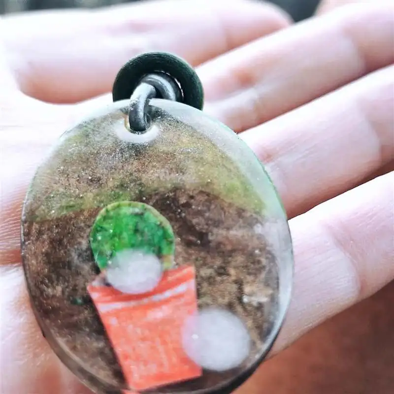 Colgante artesanal de resina con una pequeña maceta y planta en su interior, sostenido en la palma de la mano